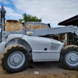 Szary w&oacute;zek widłowy teleskopowy Manitou, częściowo zabezpieczony taśmą malarską, po świeżym malowaniu, widoczny na zewnątrz w otoczeniu drzew i budynku.