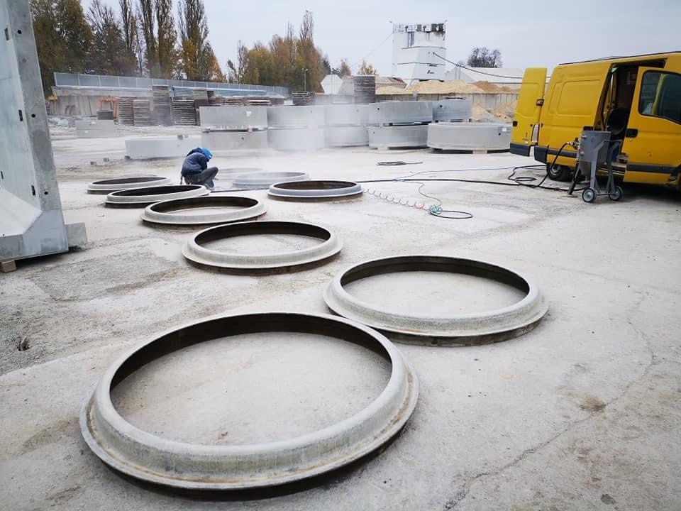 Pracownik w niebieskim kombinezonie czyści powierzchnię betonowych pierścieni na zewnątrz, obok żółtego busa z urządzeniem do obróbki strumieniowo-ściernej.