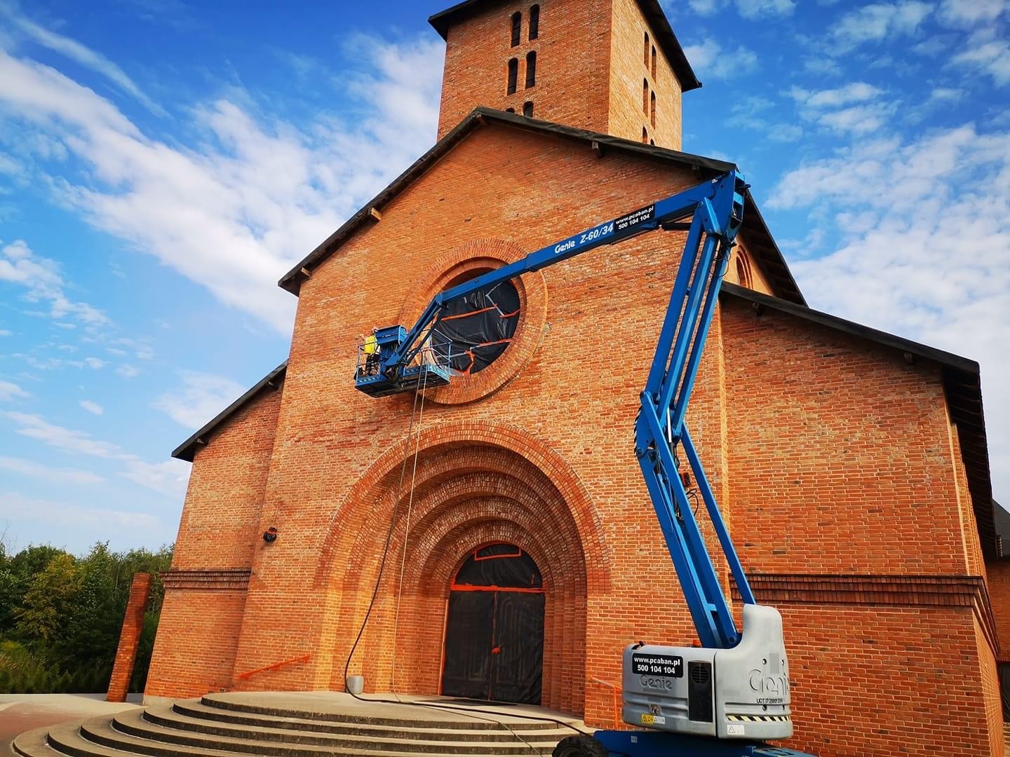 Piaskowanie ceglanej elewacji kościoła z użyciem podnośnika koszowego Genie Z-60/34, widoczne zabezpieczenie okna folią.