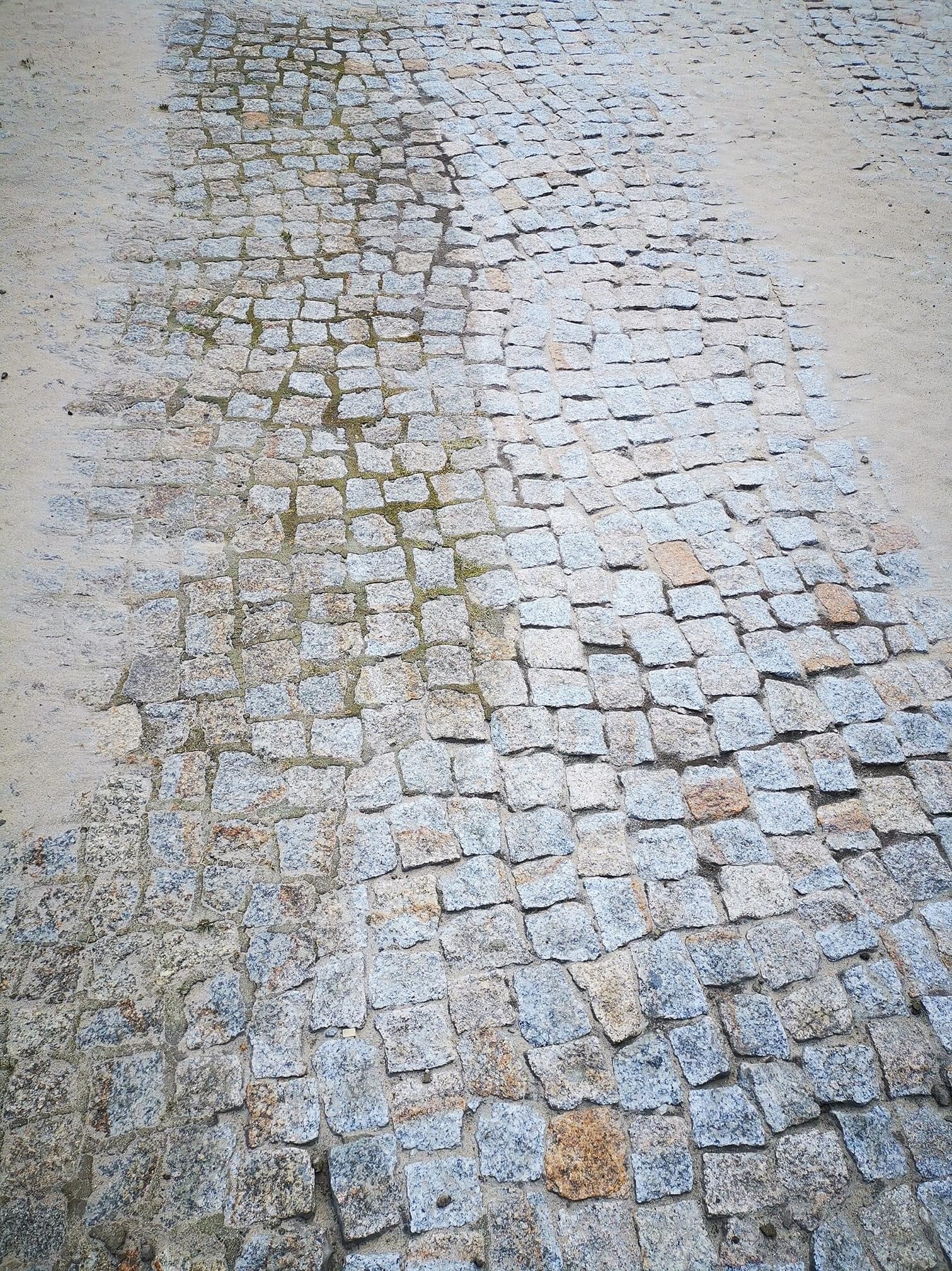 Ułożona z kostki brukowej, mokra nawierzchnia drogi z widocznymi śladami piasku po bokach.