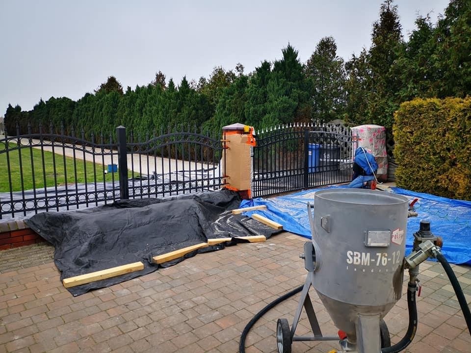 Przygotowanie do obróbki metalowej bramy: zabezpieczona folią i kartonem, widoczna piaskarka SBM-76-12 na brukowanym podjeździe, pracownik w tle.