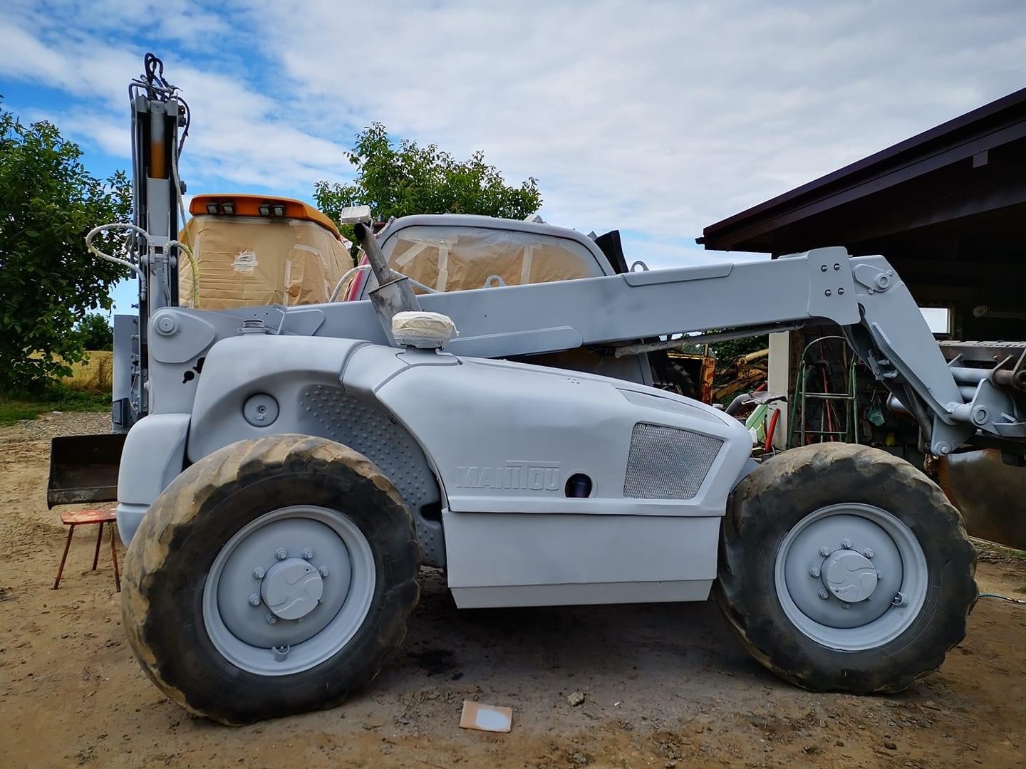 Szary wózek widłowy teleskopowy Manitou, częściowo zabezpieczony taśmą malarską, po świeżym malowaniu, widoczny na zewnątrz w otoczeniu drzew i budynku.