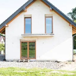 Surowy stan domu jednorodzinnego z białego bloczka, widoczne okna z brązowymi ramami, dach pokryty czarną dachówką, teren wokół domu częściowo zasypany gruzem, w tle zieleń drzew i budynek...