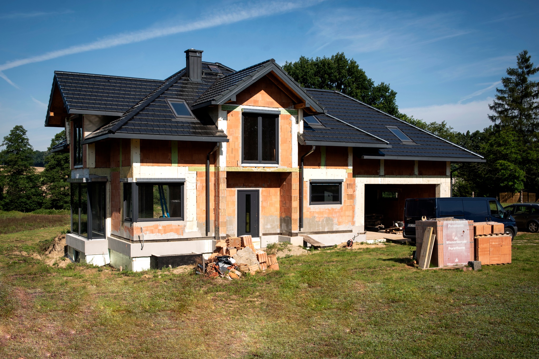 Dom jednorodzinny w trakcie budowy: ściany z czerwonej cegły Porotherm, czarny dach z dachówką, czarne okna i drzwi, widoczny stos cegieł na trawniku, samochód dostawczy w tle.