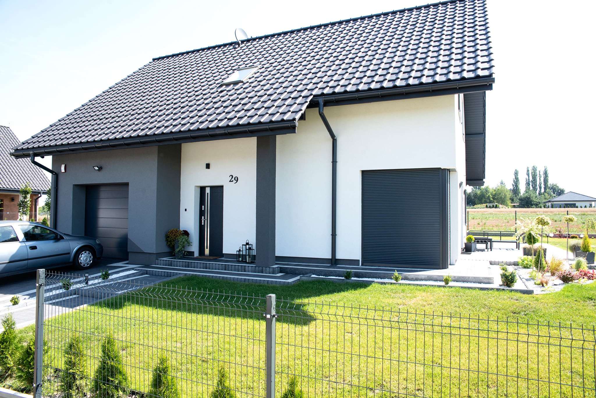 Nowoczesny dom jednorodzinny z szarą bramą garażową roletową, ciemną stolarką okienną i drzwiową oraz zielonym ogrodzeniem panelowym na pierwszym planie.
