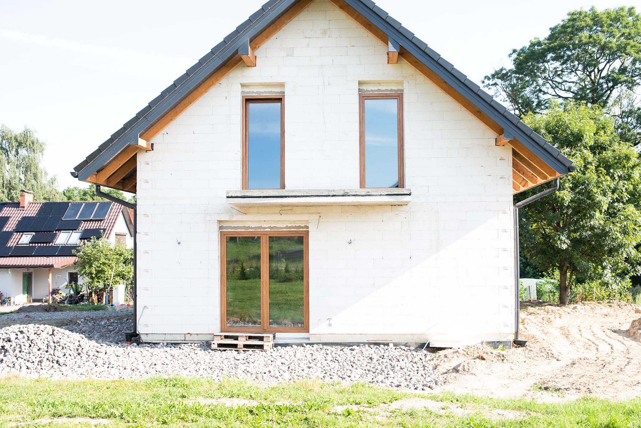 Surowy stan domu jednorodzinnego z białego bloczka, widoczne okna z brązowymi ramami, dach pokryty czarną dachówką, teren wokół domu częściowo zasypany gruzem, w tle zieleń drzew i budynek...