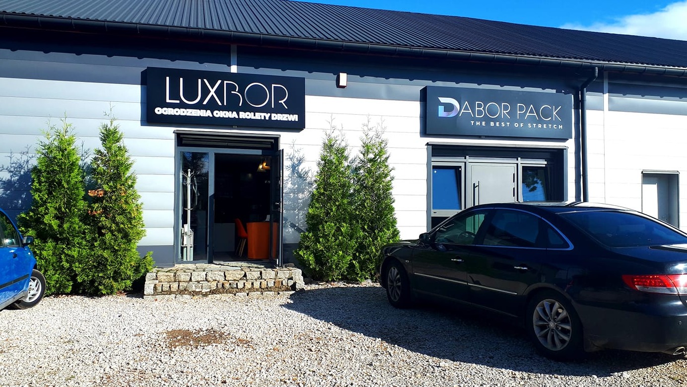 Budynek firmy LUXBOR z szyldem informującym o ofercie: ogrodzenia, okna, rolety, drzwi, widok z zewnątrz, na pierwszym planie szara nawierzchnia z kamyczków i zaparkowane samochody.