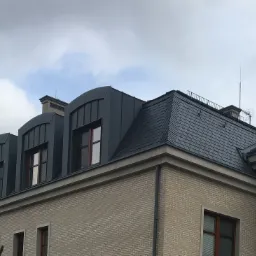 Dach z ciemnej dachówki łupkowej, z trzema lukarnami krytymi blachą, na budynku z jasnej cegły.