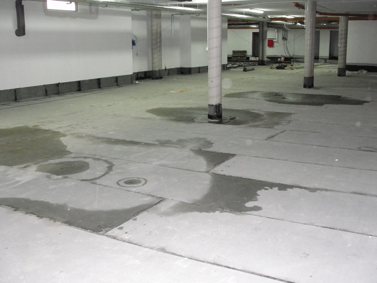 Zalana posadzka w piwnicy z widocznymi plamami wody na płytach betonowych, wsparta na kolumnach, z widocznymi rurami pod sufitem.