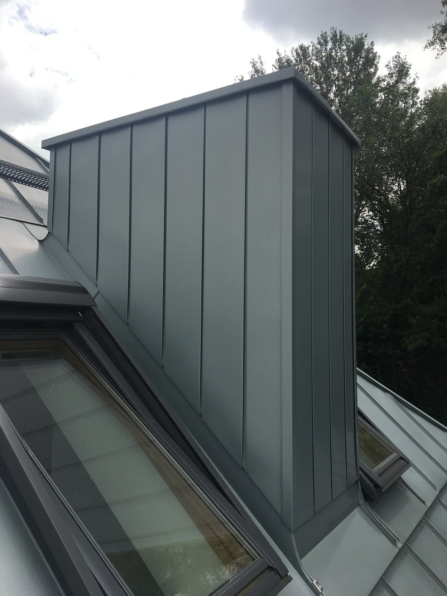 Szary komin obłożony panelami na dachu z oknami dachowymi, widok z dołu na tle zieleni.