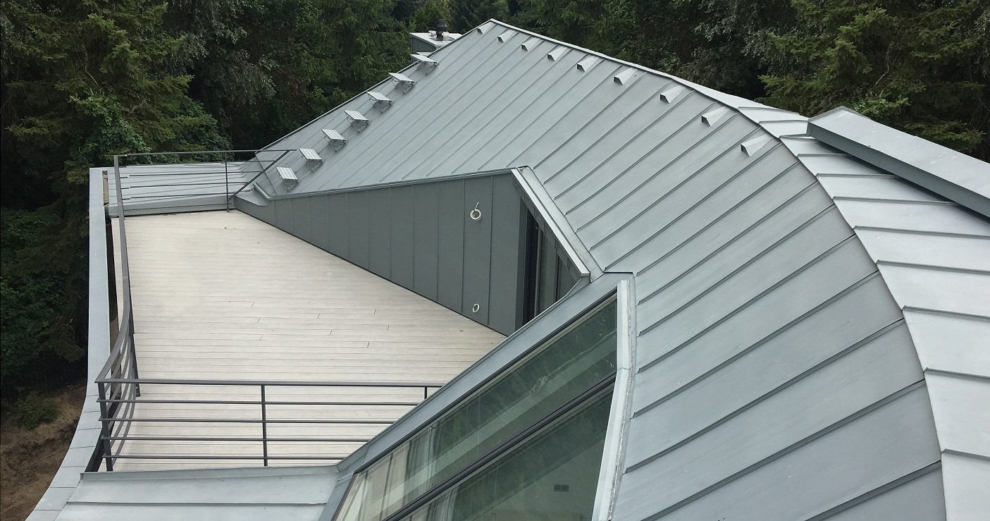 Nowoczesny dach z blachy trapezowej w odcieniach szarości, z elementami wykończeniowymi i balustradą na tle zieleni drzew.