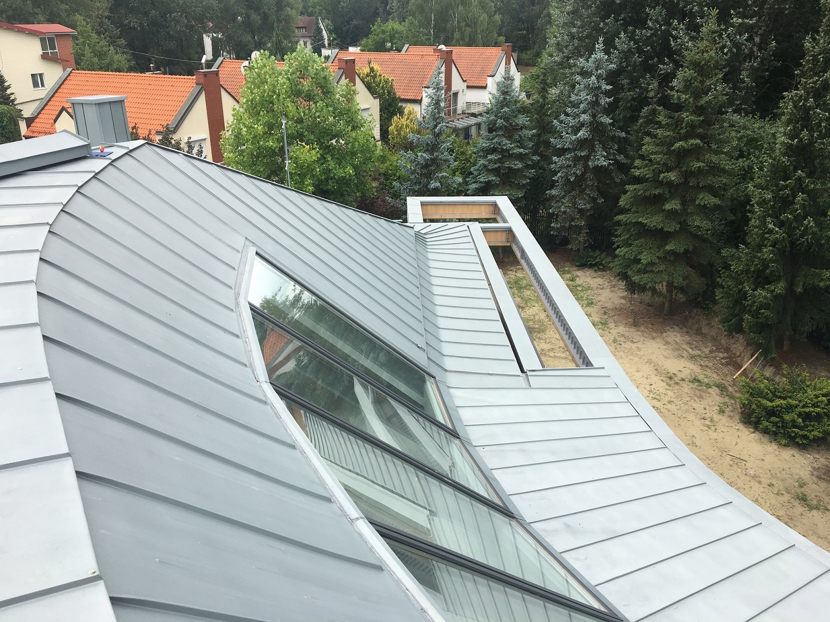 Szary dach z blachy trapezowej z oknami dachowymi i elementami konstrukcyjnymi widocznymi w otworze, w tle domy z czerwonymi dachami i drzewa.