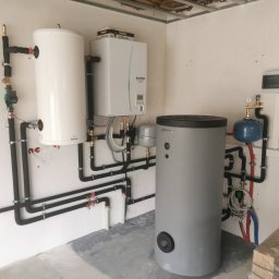 Hydrosun Sp. z o.o. - Nowo zainstalowany system z białym modułem Ecodan, dwoma pionowymi zbiornikami (Galmet, Atrinity) i czarnymi, izolowanymi rurami w pomieszczeniu technicznym.