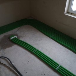 Hydrosun Sp. z o.o. - Etap montażu zielonych rur instalacyjnych na surowej betonowej posadzce, z widocznym białym rozdzielaczem i oknem z pianką montażową.