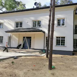 Dwupiętrowy dom z białą elewacją i ciemnym dachem, widoczny mężczyzna przechodzący obok oraz drabina przy wejściu.