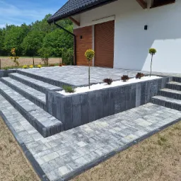 Wykończony taras i schody wejściowe do domu, wyłożone szaro-białą kostką brukową, z ozdobnymi rabatami wypełnionymi białym kamieniem i niskimi drzewkami.