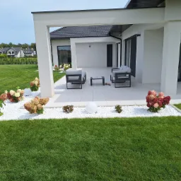 Nowoczesny taras z białymi płytami, minimalistyczne meble ogrodowe, rabaty z białym kamieniem i hortensjami w odcieniach różu i zieleni, zadbany trawnik.