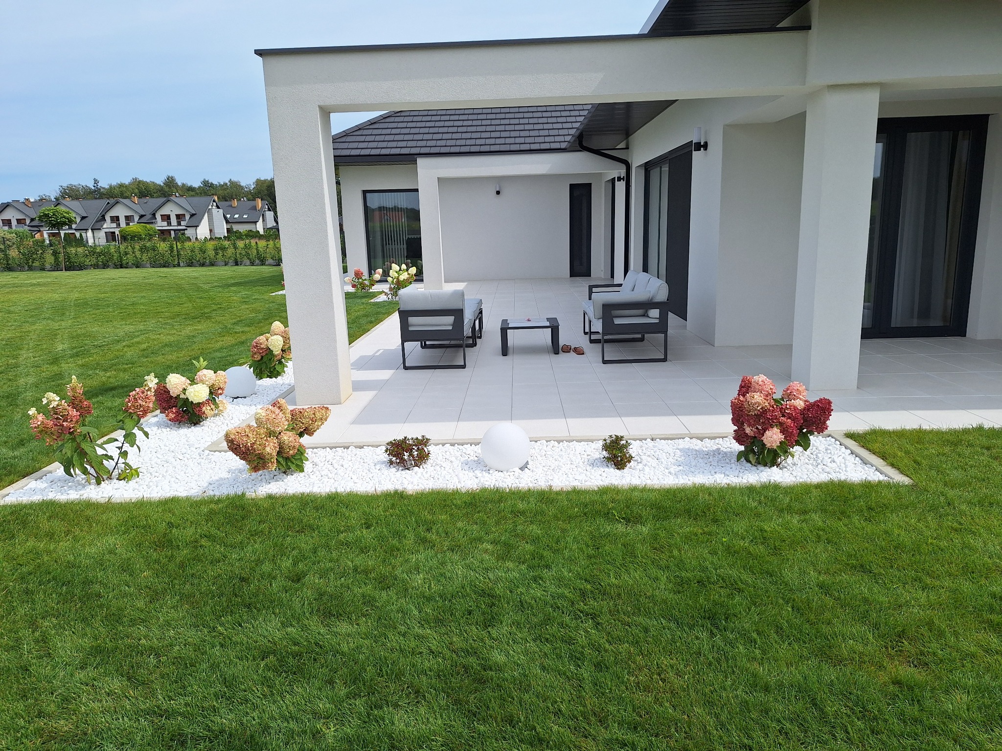 Nowoczesny taras z białymi płytami, minimalistyczne meble ogrodowe, rabaty z białym kamieniem i hortensjami w odcieniach różu i zieleni, zadbany trawnik.