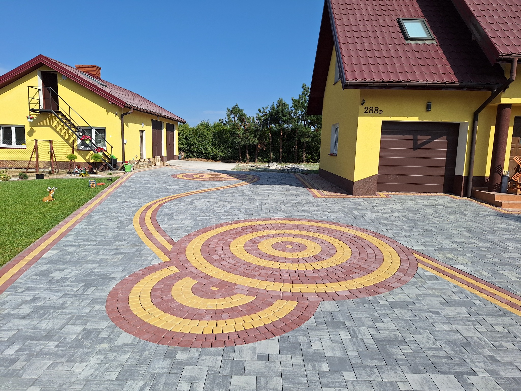 Ogród z dwoma budynkami i ozdobnym podjazdem z szarej i kolorowej kostki brukowej ułożonej we wzór koncentrycznych okręgów i linii.