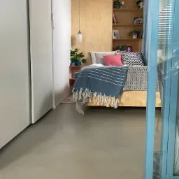 Jasnoszara podłoga w sypialni z łóżkiem i zabudowaną półką, widok zza błękitnych drzwi.