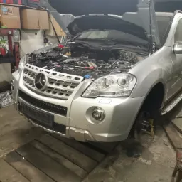 Srebrny samochód z otwartą maską w warsztacie, brak koła, widoczny silnik i elementy zawieszenia.