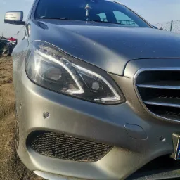Przednia część szarego samochodu marki Mercedes z widoczną lampą, grillem i czujnikami parkowania, lekko zabrudzona, na tle ziemi i błękitnego nieba.