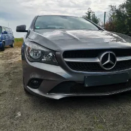 Szary Mercedes CLA z widocznym przodem, grillem i logo na tle pochmurnego nieba i zieleni.