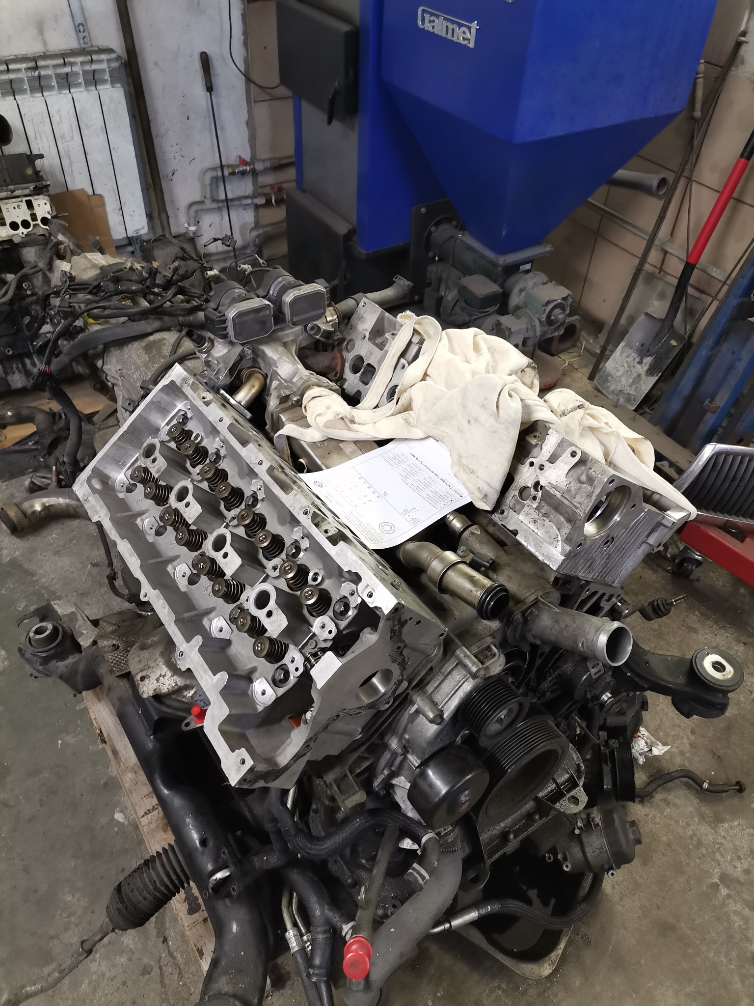 Rozłożony silnik samochodowy z widoczną głowicą cylindrów, blokiem silnika, przewodami i innymi częściami, leżący na palecie w warsztacie.