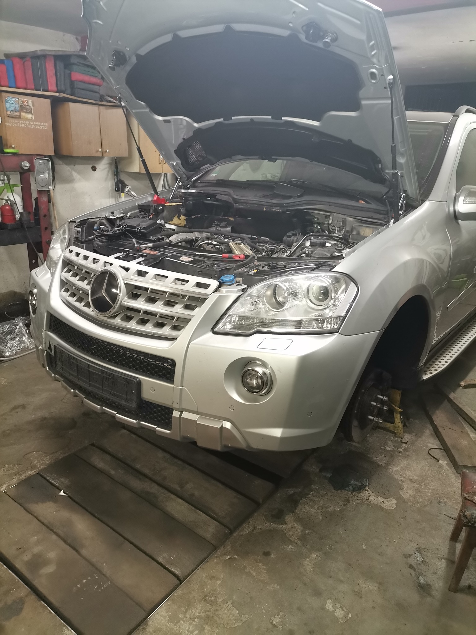 Srebrny samochód z otwartą maską w warsztacie, brak koła, widoczny silnik i elementy zawieszenia.