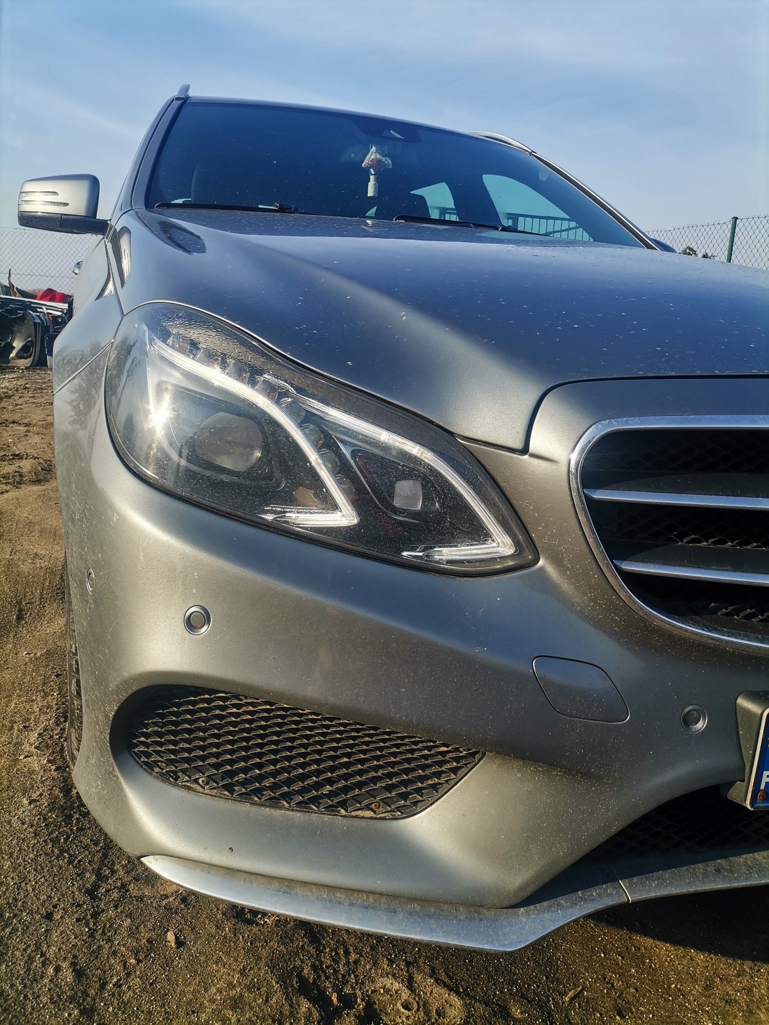 Przednia część szarego samochodu marki Mercedes z widoczną lampą, grillem i czujnikami parkowania, lekko zabrudzona, na tle ziemi i błękitnego nieba.