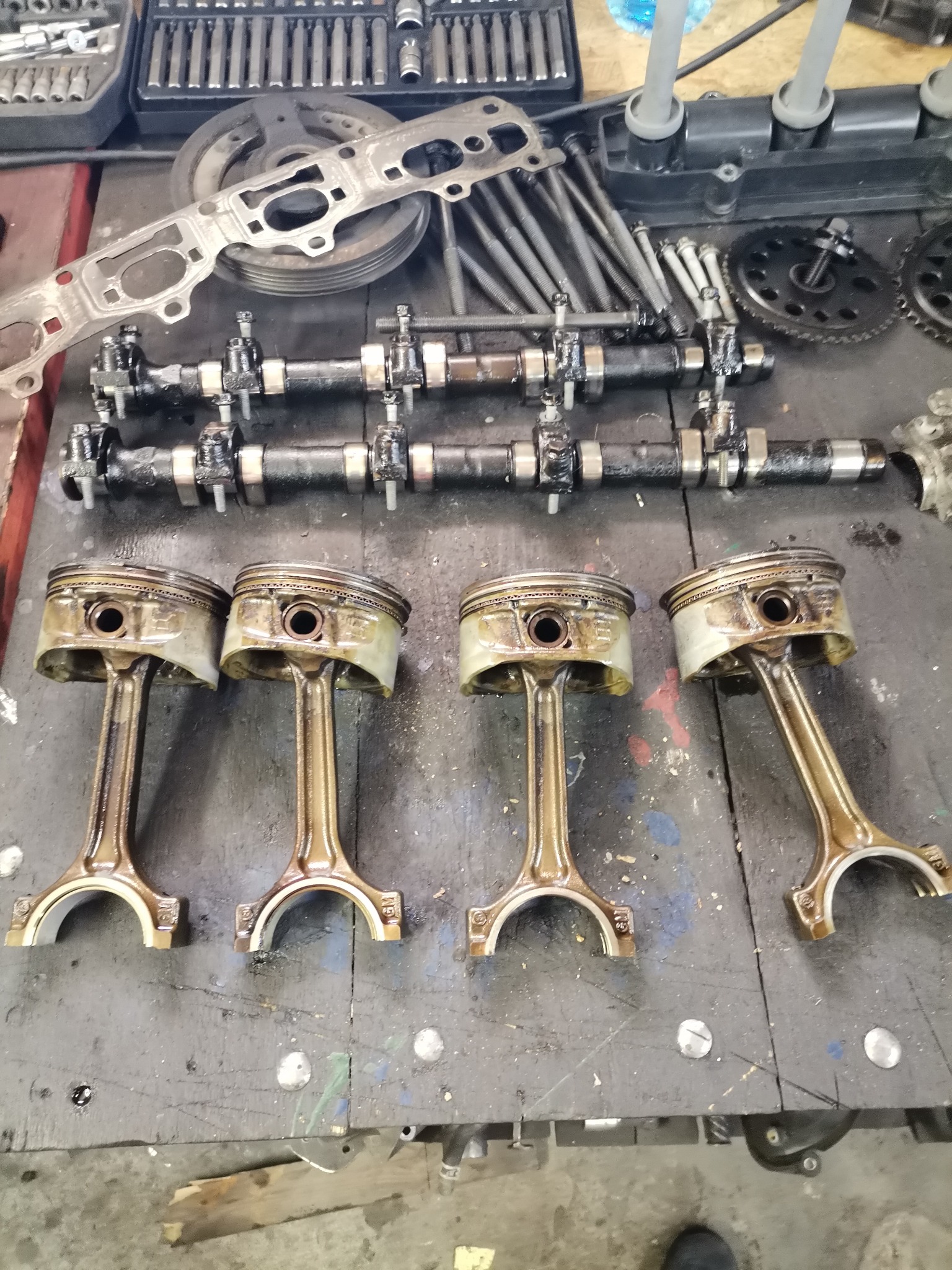 Cztery zużyte tłoki silnikowe ułożone obok siebie na brudnym warsztatowym stole, powyżej wałki rozrządu, klucze nasadowe i inne części samochodowe.