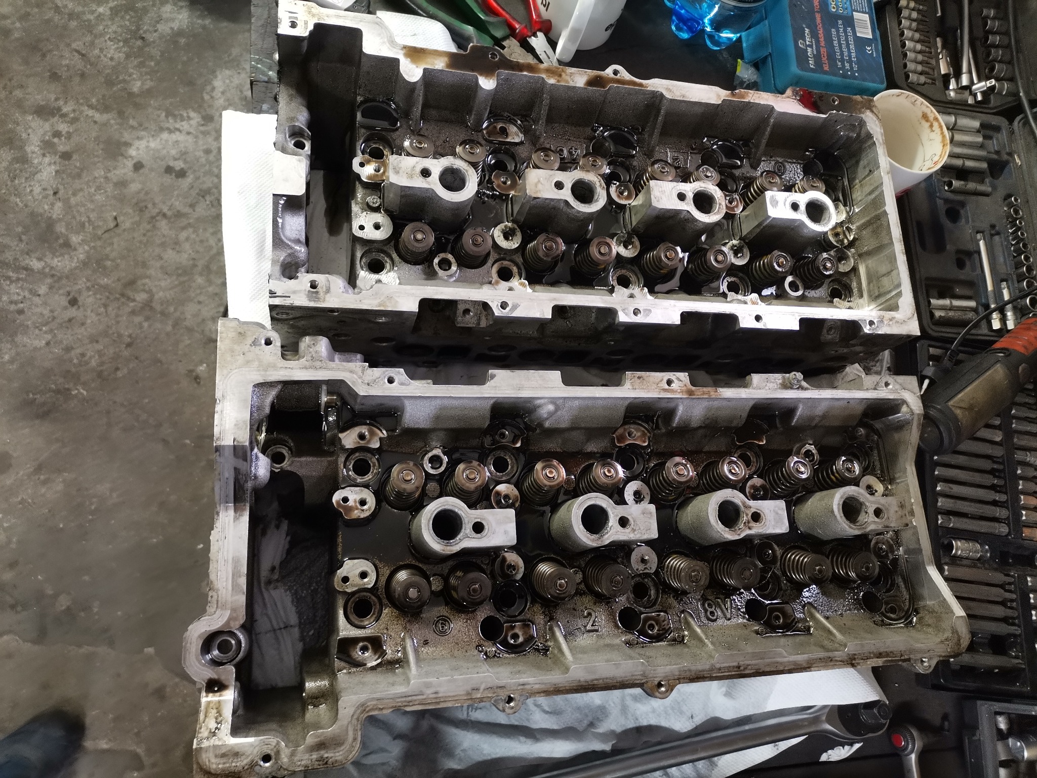 Dwie zdjęte głowice silnika spalinowego, widoczne sprężyny zaworowe i kanały olejowe, zabrudzone olejem, obok skrzynka z narzędziami i klucze nasadowe
