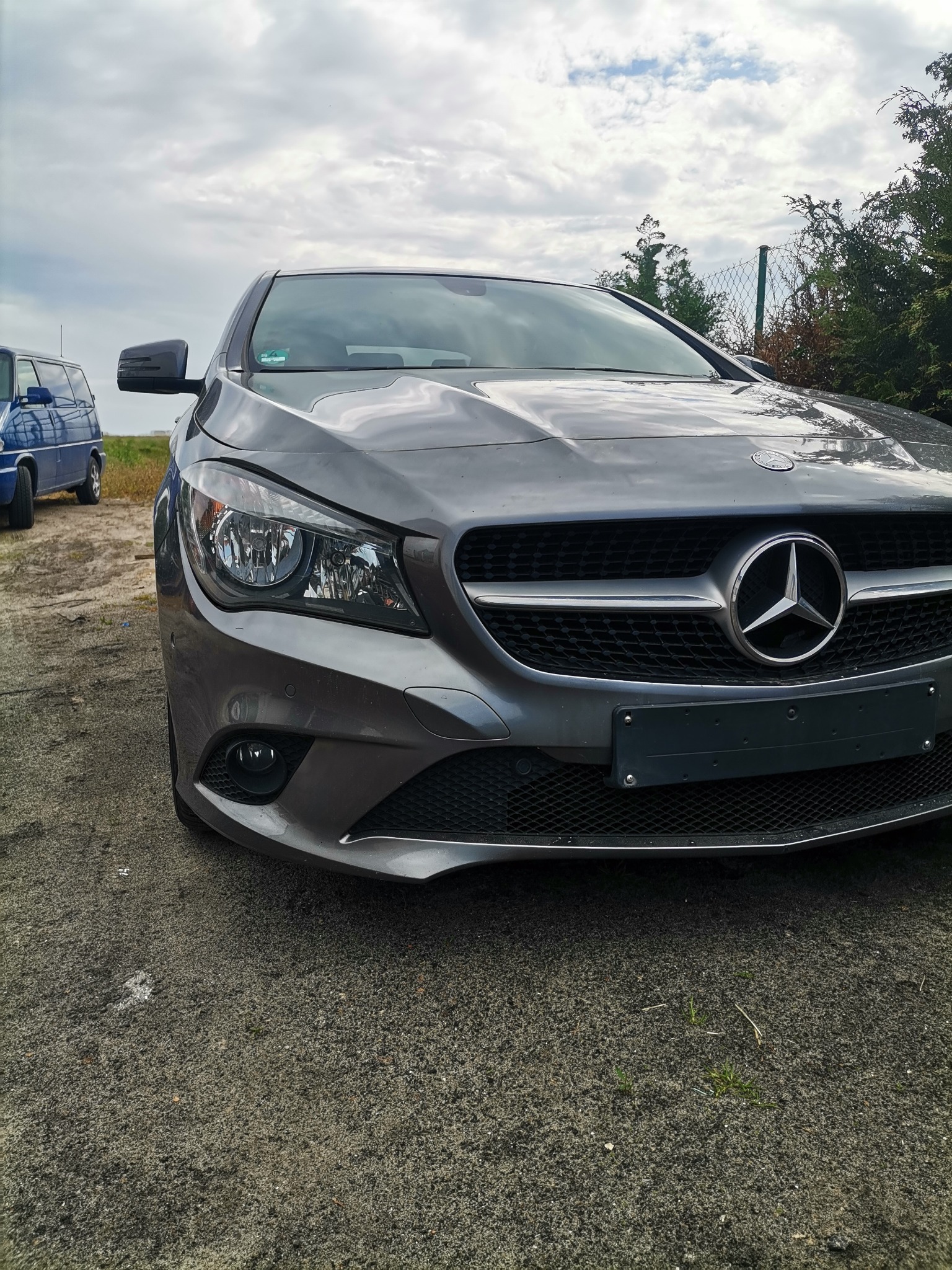Szary Mercedes CLA z widocznym przodem, grillem i logo na tle pochmurnego nieba i zieleni.