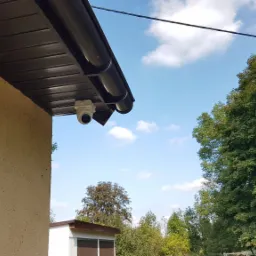 Kamera kopułkowa zamontowana pod okapem budynku z elewacją w kolorze piaskowym, na tle błękitnego nieba z chmurami i zieleni drzew.