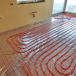 Andem - Pomarańczowe rury ogrzewania podłogowego ułożone na srebrnej folii izolacyjnej z czerwoną siatką, widoczne przyłącza na ścianie i okno w tle.