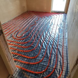 Andem - Pomarańczowe rury ogrzewania podłogowego ułożone na folii z rastrem, widok z góry, surowy stan pomieszczenia z oknem dachowym.