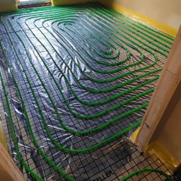 Andem - Instalacja ogrzewania podłogowego z zielonymi rurkami ułożonymi w pętle na folii z rastrem, widoczne mocowania rurek oraz fragment okna i ściany.