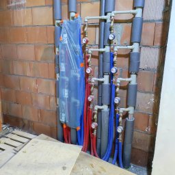 Andem - Rozdzielacz ogrzewania podłogowego w folii ochronnej, rury doprowadzające ciepło w kolorze czerwonym i powrotne w kolorze niebieskim, zaizolowane piony, widoczna surowa ściana z cegły.