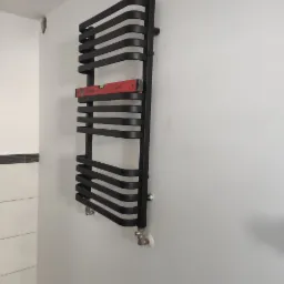 Czarny, designerski grzejnik łazienkowy drabinkowy, zamontowany na białej ścianie, poziomowany czerwoną poziomicą, widoczne przyłącza instalacji.