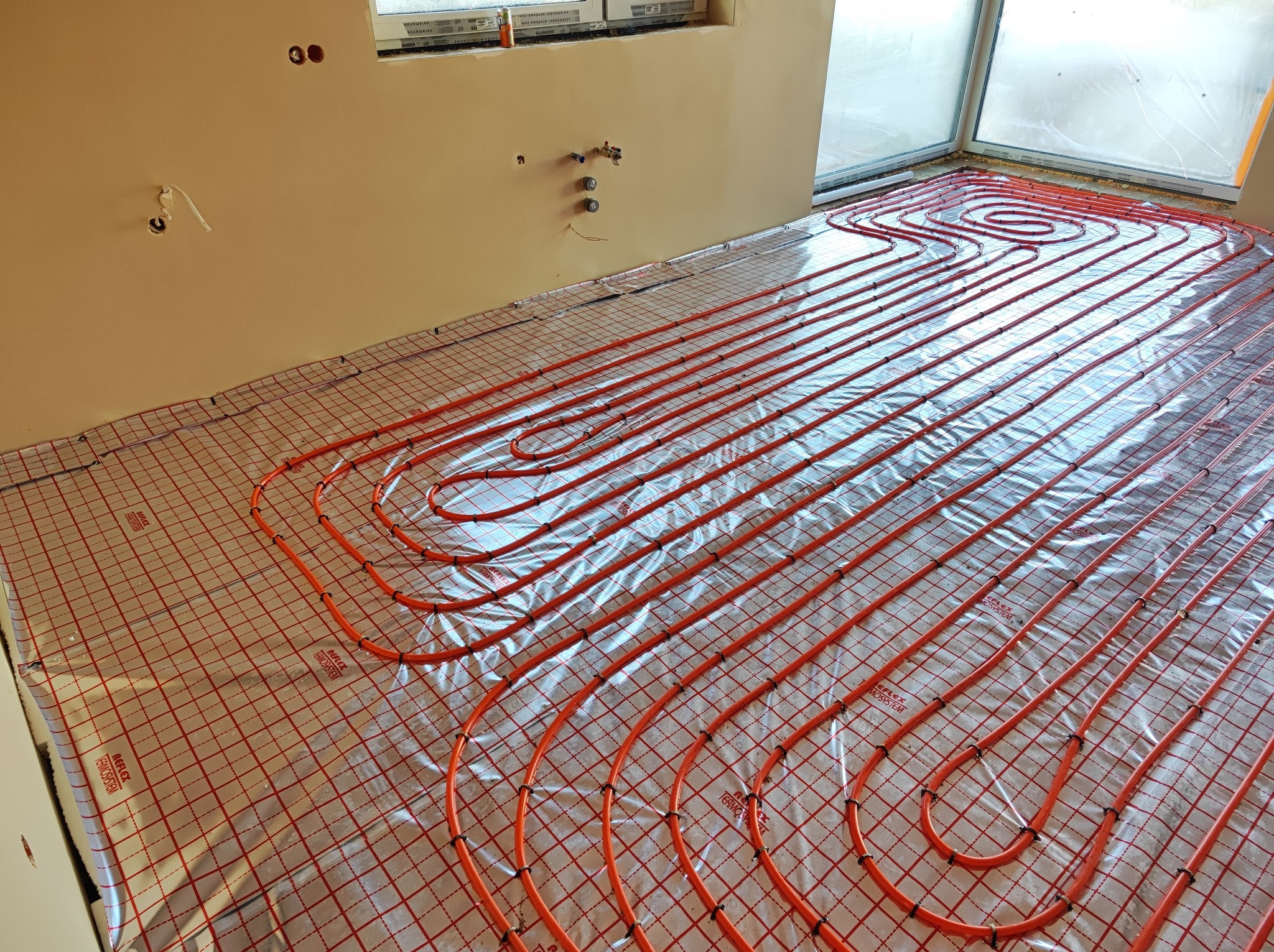 Pomarańczowe rury ogrzewania podłogowego ułożone na srebrnej folii izolacyjnej z czerwoną siatką, widoczne przyłącza na ścianie i okno w tle.