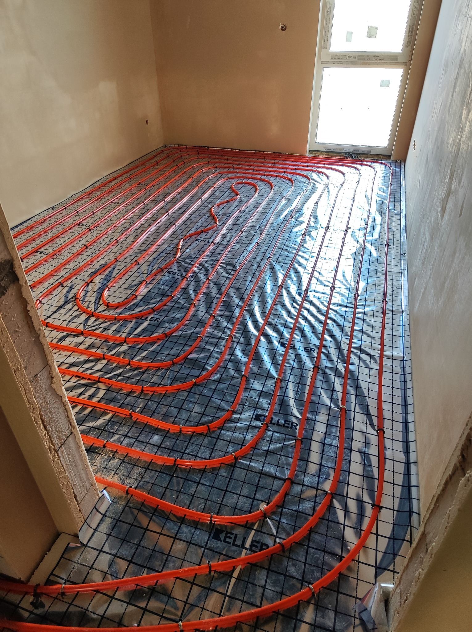 Pomarańczowe rury ogrzewania podłogowego ułożone na folii z rastrem, widok z góry, surowy stan pomieszczenia z oknem dachowym.