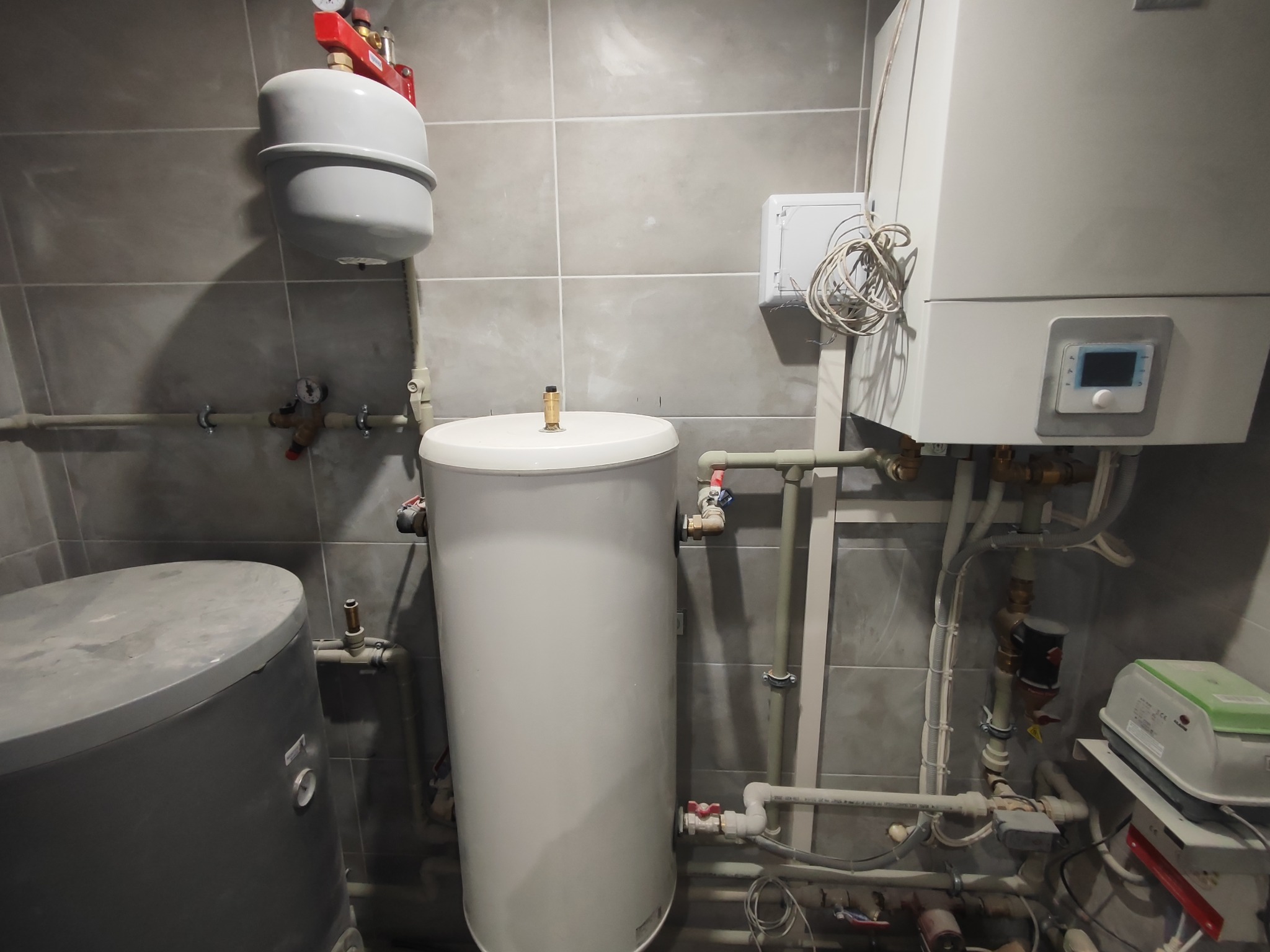 Wnętrze kotłowni z zasobnikiem ciepłej wody, kotłem gazowym, naczyniem przeponowym i instalacją hydrauliczną na tle szarych płytek.