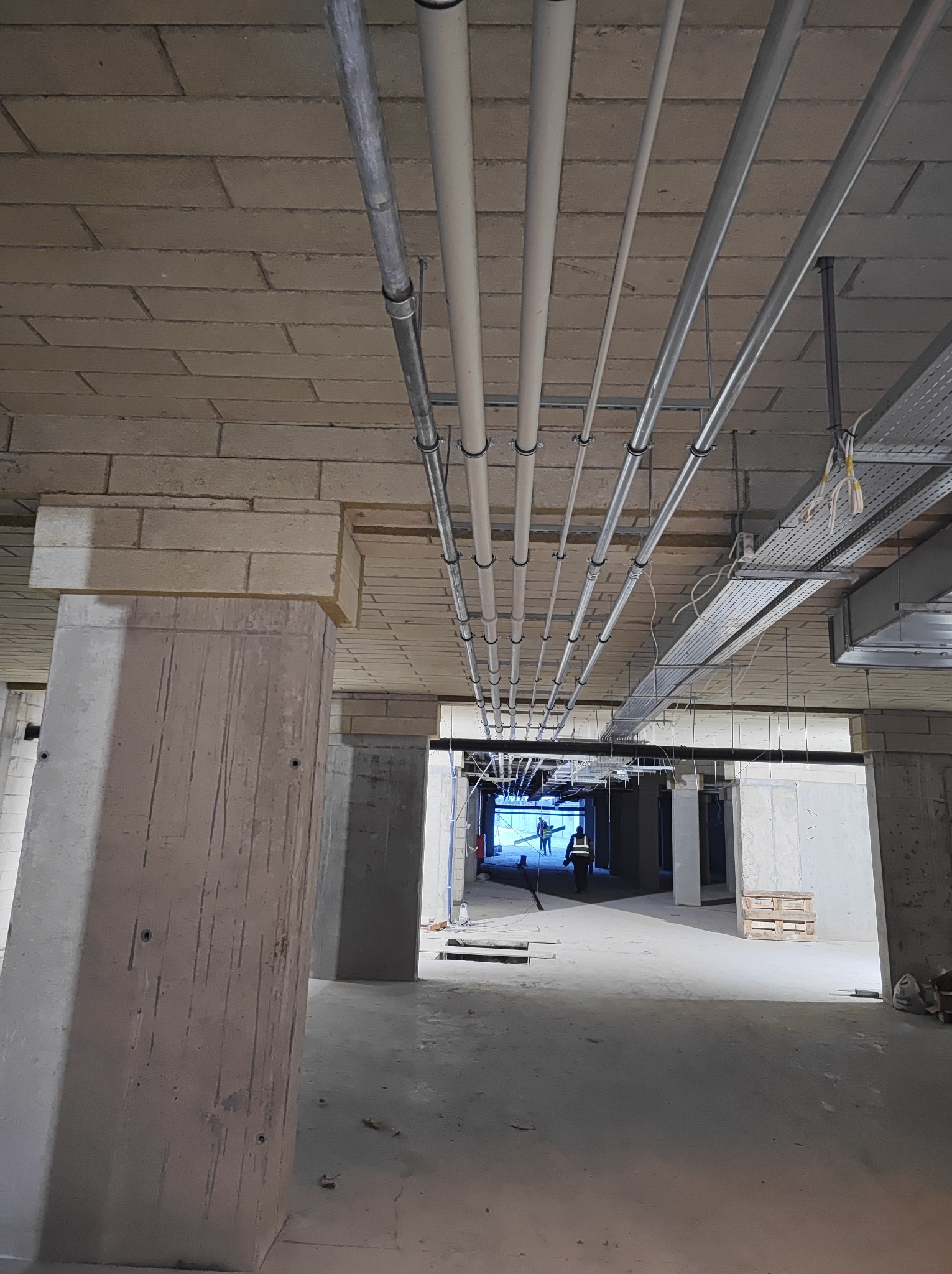 Surowy betonowy sufit z rzędami rur i przewodów instalacyjnych, widoczne elementy konstrukcyjne budynku, w tle sylwetki pracowników.