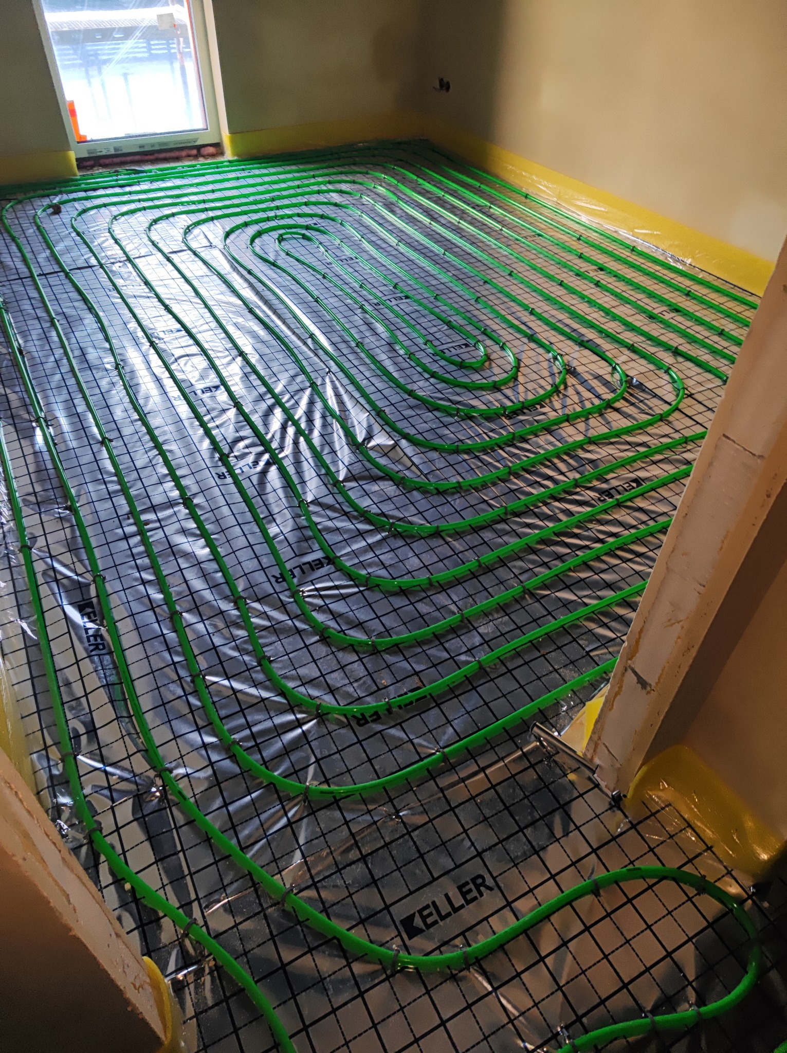 Instalacja ogrzewania podłogowego z zielonymi rurkami ułożonymi w pętle na folii z rastrem, widoczne mocowania rurek oraz fragment okna i ściany.