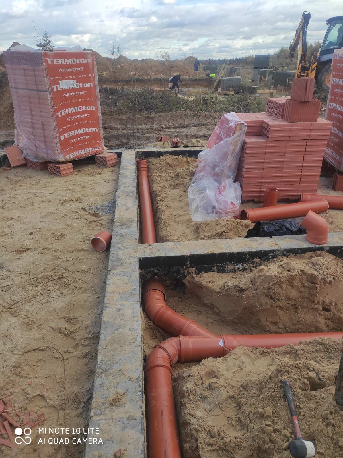 Wykonane wykopy pod fundamenty z widocznymi rurami kanalizacyjnymi w piasku, palety z cegłami w tle i koparka na budowie.