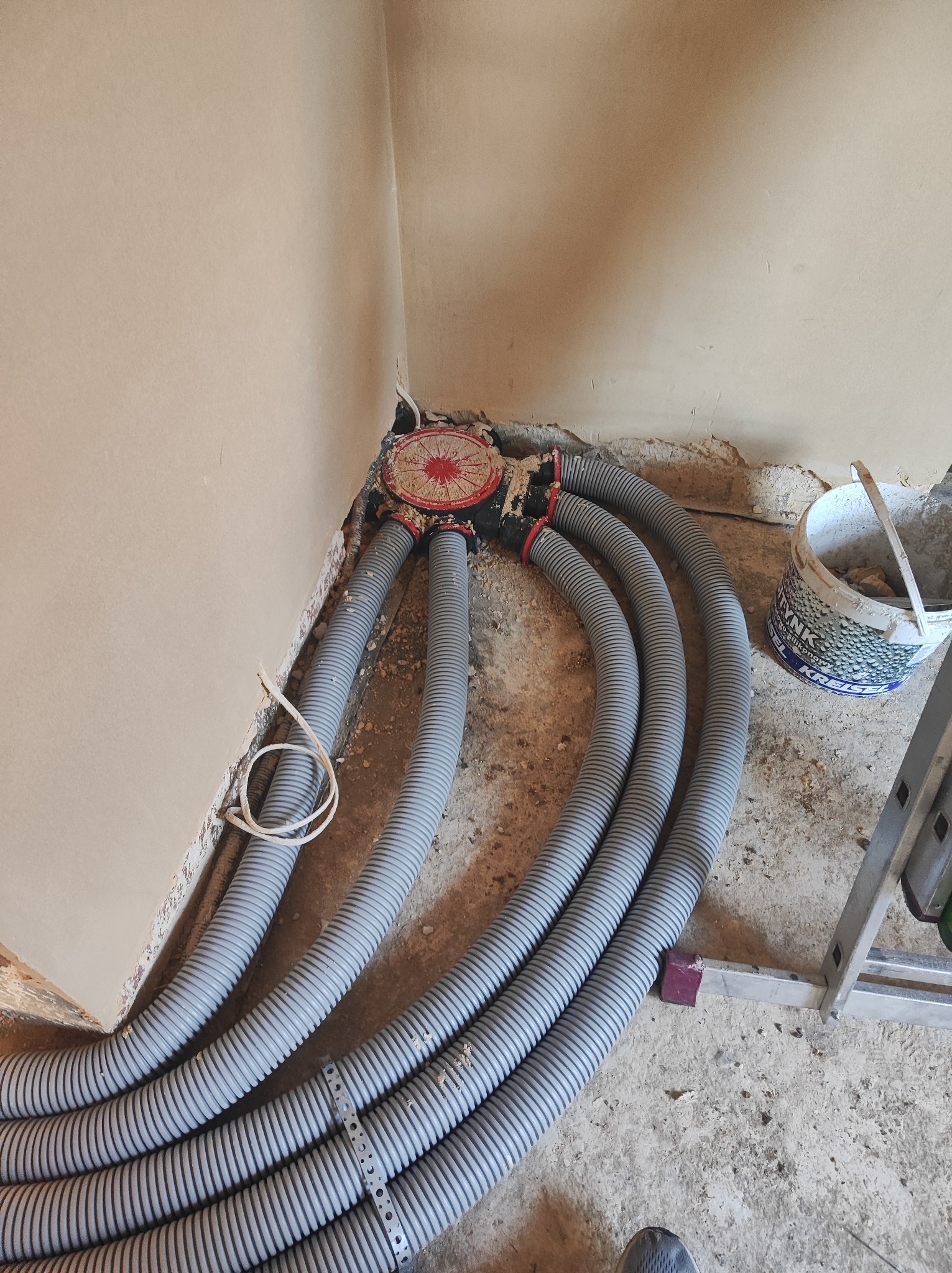 Szare, karbowane rury instalacyjne wychodzące z rozdzielacza w rogu pomieszczenia, ściany w stanie surowym, widoczne ślady po kuciu, obok stoi wiadro z zaprawą.