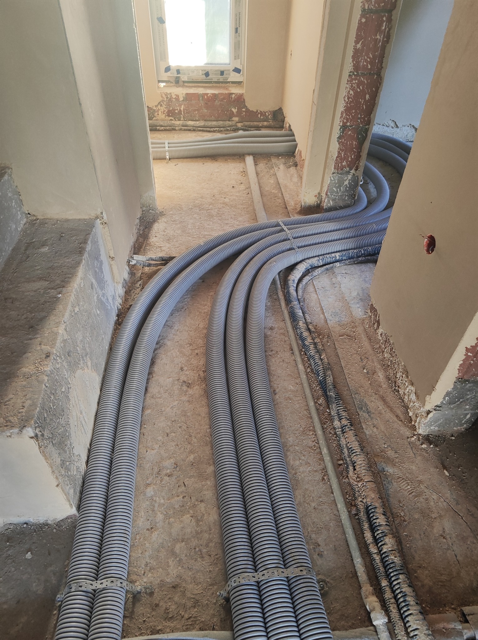 Szereg szarych, karbowanych rur instalacyjnych ułożonych równolegle na podłodze w trakcie remontu, zabezpieczonych metalowymi uchwytami, z widocznymi śladami zaprawy i niedokończonymi ścianami.