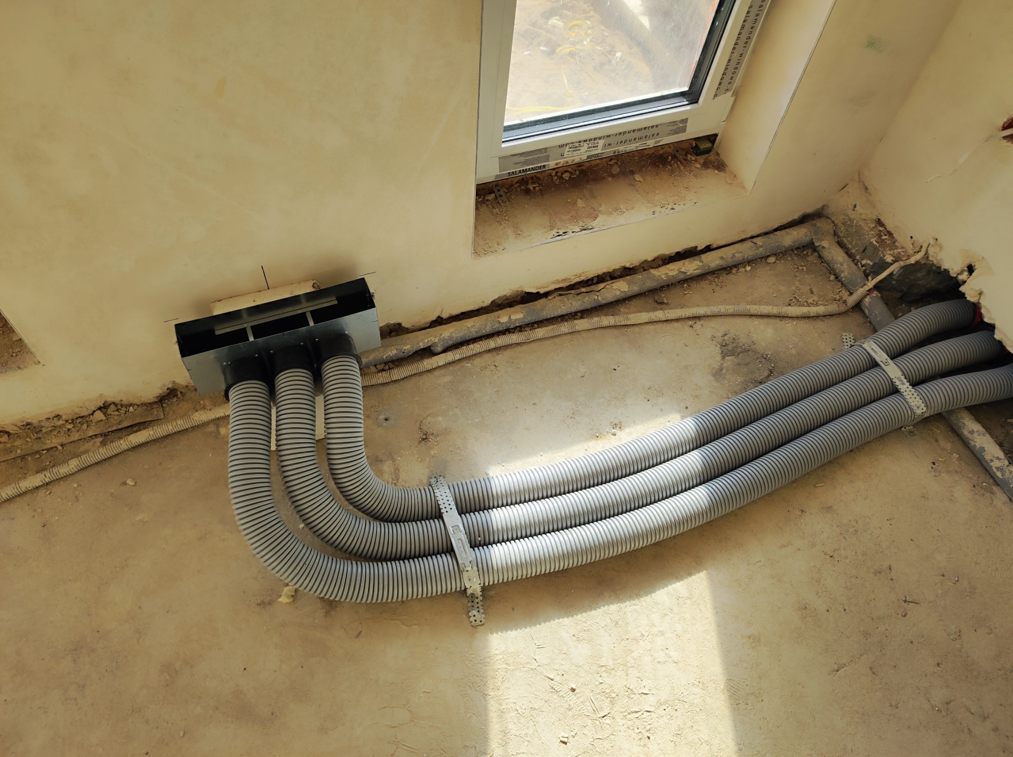 Trzy szare, karbowane rury instalacyjne wychodzące ze ściany w surowym stanie, zabezpieczone uchwytem, widoczne przyłącza i okno w tle.