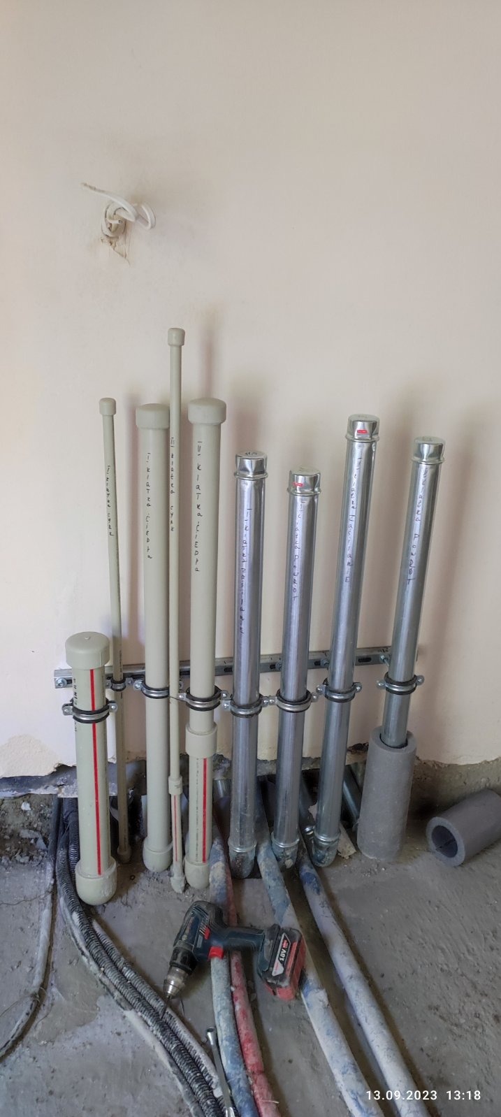 Instalacja pionów wodnych i kanalizacyjnych z rur PP i stalowych, zamocowanych do ściany za pomocą obejm i szyn montażowych, w tle widoczne przewody elektryczne w peszlach i wiertarka akumulatorowa.
