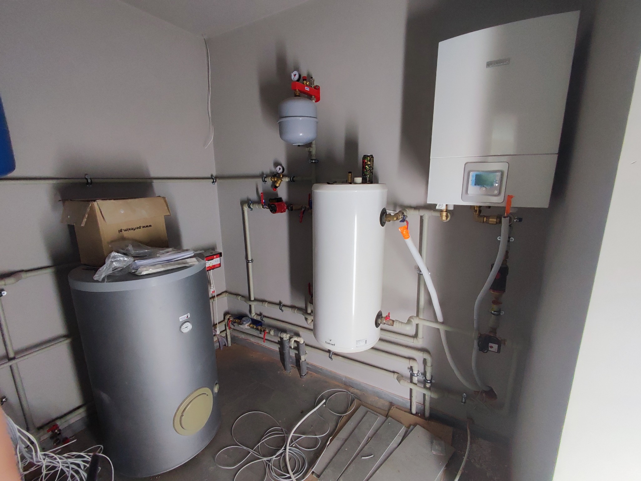 Kotłownia w trakcie montażu: widoczne zbiorniki, piec gazowy, rury instalacyjne z zaworami, manometry oraz rozłożone przewody na podłodze.